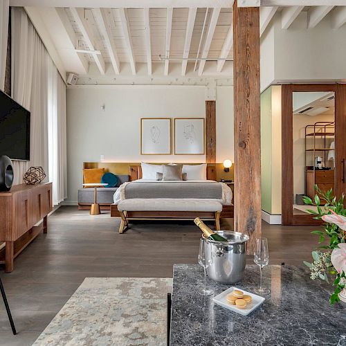 A modern, stylish hotel room with a bed, TV, and a seating area featuring a marble table with flowers, a champagne bucket, and two glasses.