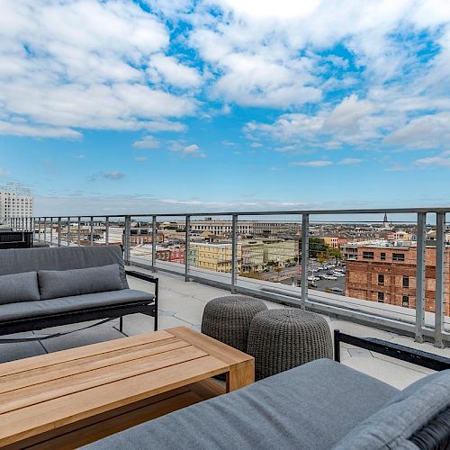 The image shows a rooftop patio with modern outdoor furniture, a table, and a cityscape view under a partly cloudy sky, surrounded by a glass railing.