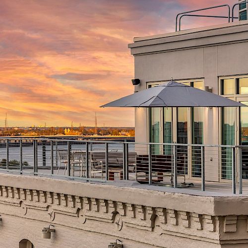 A rooftop patio with a metal railing, outdoor seating, and an umbrella, overlooking a cityscape and river at sunset.