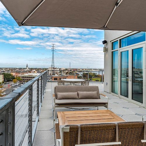 A rooftop patio with modern outdoor furniture overlooks a cityscape under a partly cloudy sky. Large windows and an umbrella are visible.