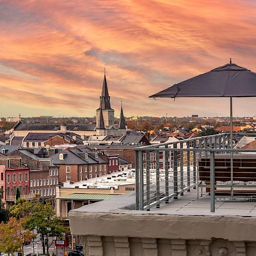 A rooftop terrace with a lounge chair and umbrella overlooks a cityscape with colorful buildings and a church spire under a vibrant sunset sky.