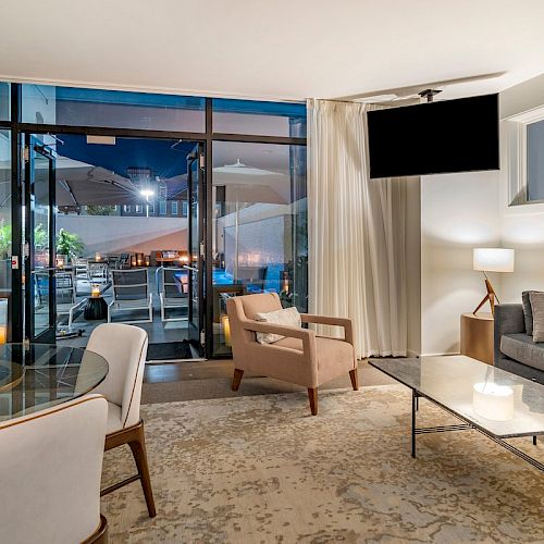 A modern living room with a glass wall opens to a patio, featuring a sofa, armchairs, a coffee table, and a view of outdoor seating and umbrellas.