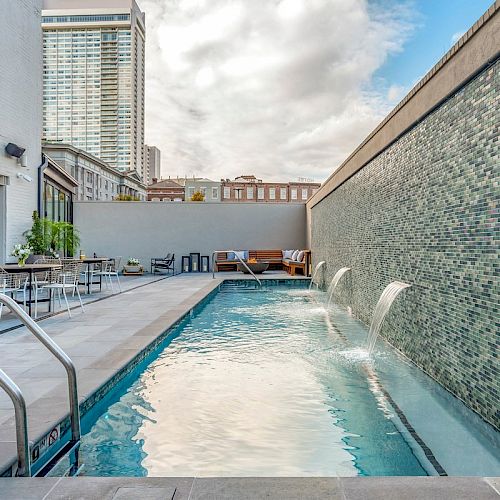 An outdoor pool with water features beside a tiled wall, lounge chairs, and tables. A building and skyscrapers are in the background.