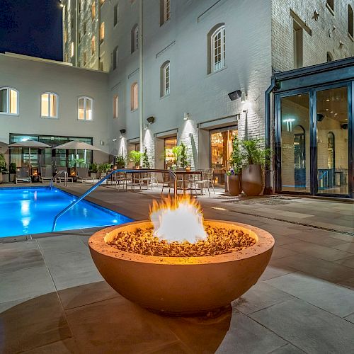 A modern outdoor patio with a large fire pit, seating, a pool, and nearby buildings lit up at night, creating a cozy ambiance.