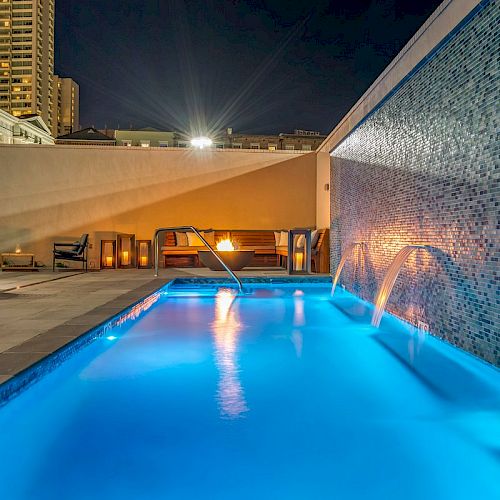 A lit pool at night with cascading waterfalls from a wall, lounge chairs, a fire pit, and a tall building in the background ending the sentence.