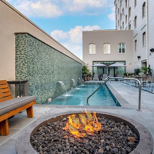 An outdoor area features a pool, fire pit, wooden bench with cushions, and tables with chairs, set against a building with large windows.