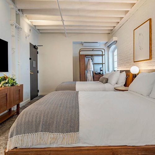 The image shows a modern hotel room with two double beds, a large TV, a wooden cabinet, and a minimalist decor including a framed art piece.