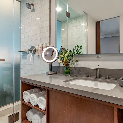 This image shows a modern bathroom with a sink, a mirror, rolled towels on shelves, and a walk-in shower with a glass door, ending the sentence.