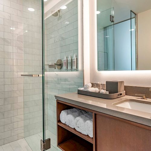 Modern bathroom with a glass-enclosed shower, tiled walls, illuminated mirror, sink with toiletries, and wooden vanity with rolled towels.