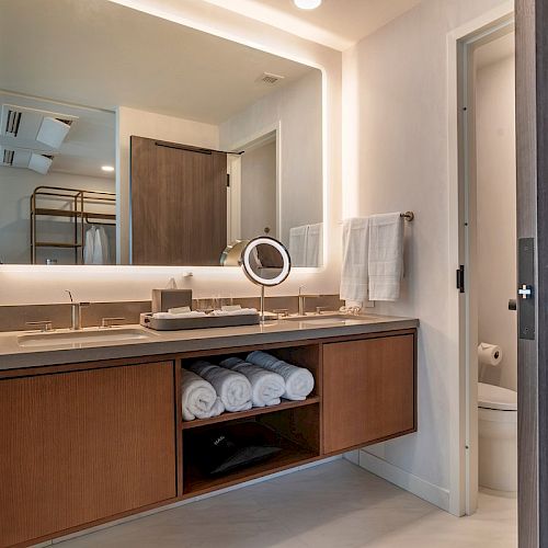 The image shows a modern bathroom with a double sink, a large mirror, towels, and a separate toilet area to the right.