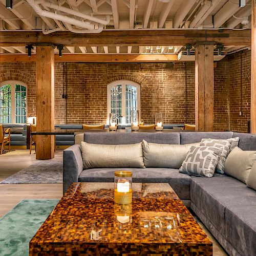 This image shows a stylish lounge with a large gray sectional sofa, wooden coffee table, brick walls, and exposed wooden beams, giving a cozy industrial vibe.