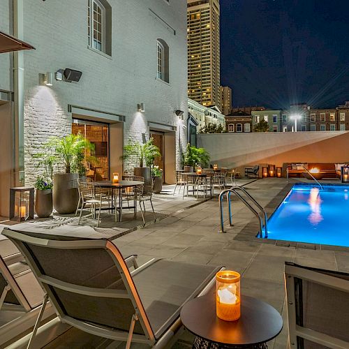 A modern outdoor pool area at night with lounge chairs, tables, candles, and plants. The pool is lit up, and buildings are visible in the background.