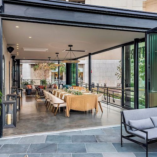 An elegant indoor-outdoor dining area with a long table set for a meal, surrounded by modern seating, glass doors, and greenery outside the seating area.