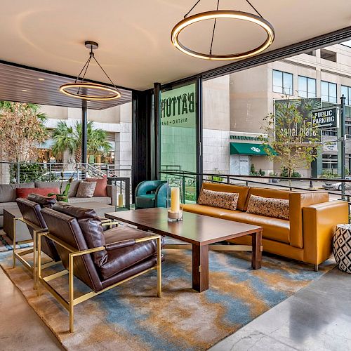 A modern lounge area with sofas, armchairs, a large table, and hanging lights, featuring a city view through floor-to-ceiling windows.