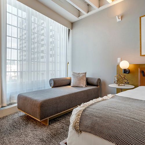 A modern bedroom with a large window, gray chaise lounge, comfortable bed with gray and white bedding, and minimalist decor on the walls.