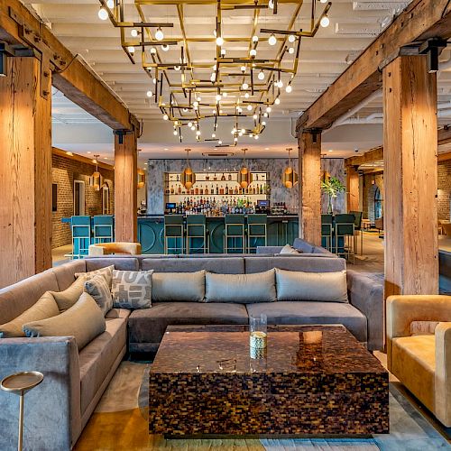 Modern lounge with exposed beams, stylish lighting, plush seating, and central coffee table; bar area behind with a chic navy-blue counter and stools.