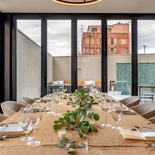 A long dining table set with glasses, plates, and greenery in a modern indoor space with large glass doors and an urban view outside.