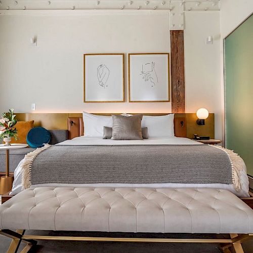 A modern bedroom featuring a plush bed with gray linens, a tufted bench, stylish wall art, bedside tables with lamps, and a minimalist decor aesthetic.