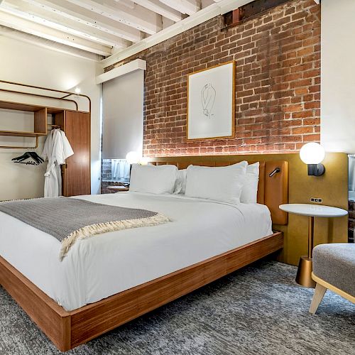 The image shows a modern, cozy bedroom with a double bed, wooden furniture, exposed brick wall, and a sitting area with a grey armchair and side table.