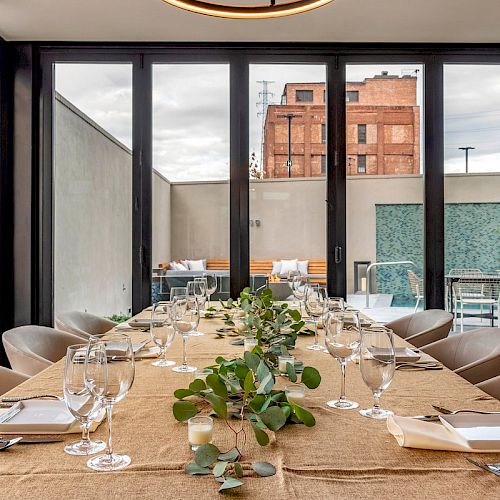 A long dining table set with glasses, plates, and greenery in a modern indoor space with large glass doors and an urban view outside.