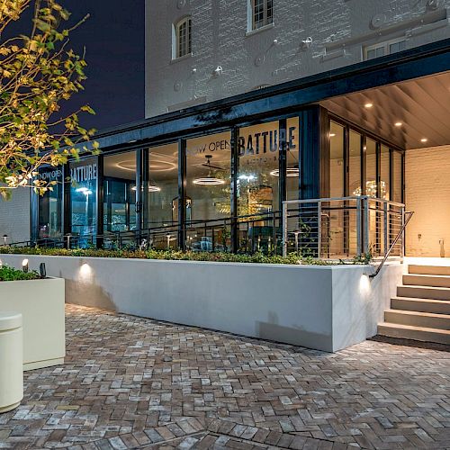 A modern cafe with large glass windows, labeled "BATTLE," is lit up at night. The entrance features a ramp and stairs, and there are plants outside.