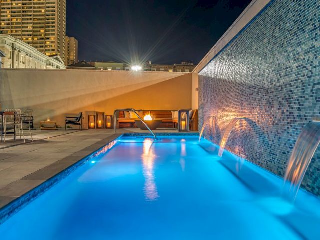 A rooftop pool with blue water and a fountain at nighttime, with a city view in the background.