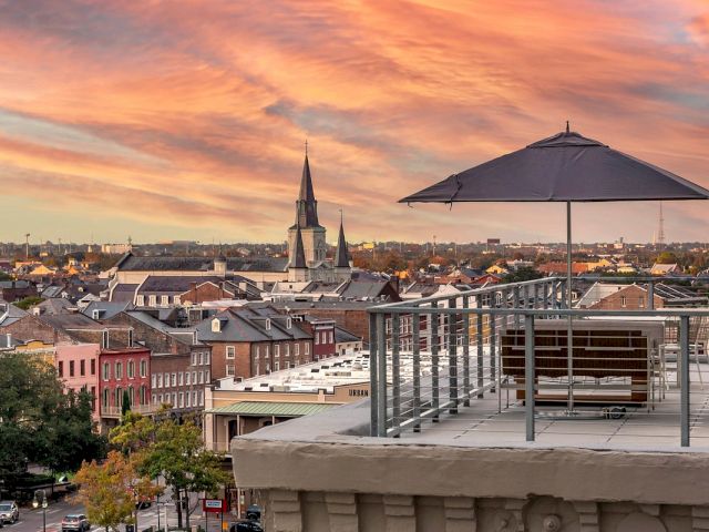 A rooftop patio with a bench, umbrella, and railing overlooking a cityscape with a church steeple at sunset.