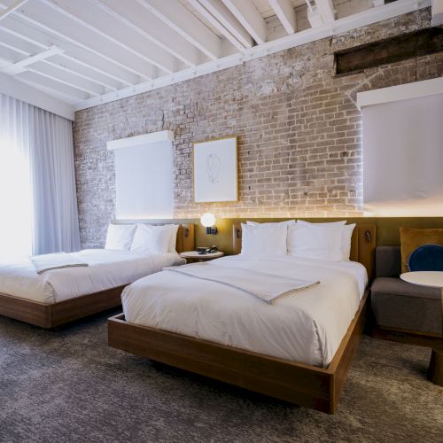 A modern hotel room with two wooden beds, white linens, brick wall, soft seating, and a bright windowed corner; stylish and airy.