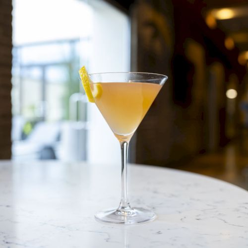 A framed cocktail in a martini glass with a lemon twist, on a marble bar near a bright, softly blurred interior backdrop.