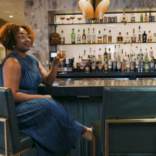 A woman with curly hair sits at a bar in a blue dress, holding a drink, with shelves full of bottles behind her.