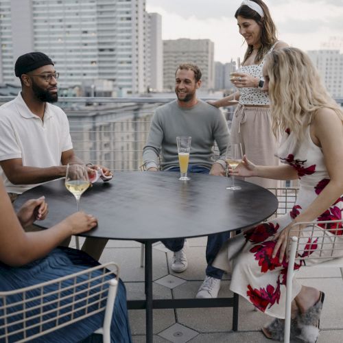 A group of five friends sits around a round outdoor table on a balcony, chatting and sharing drinks in a city setting.