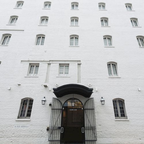 A white brick building with a black arched doorway, small barred windows, and evenly spaced tall windows rising above, all under a cloudy sky.