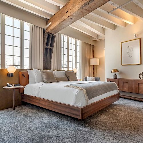 A modern bedroom with large windows, a wooden bed, neutral tones, exposed beams, abstract wall art, a bedside table, and a sideboard are depicted.