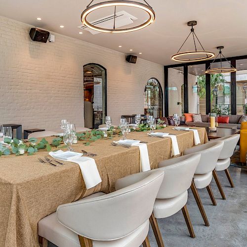 A bright, modern dining room with a long wooden table, white chairs, neutral walls, and warm pendant lighting.