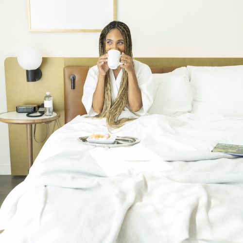 A person sits on a bed, sipping from a white cup, with a breakfast tray and bedside table nearby.