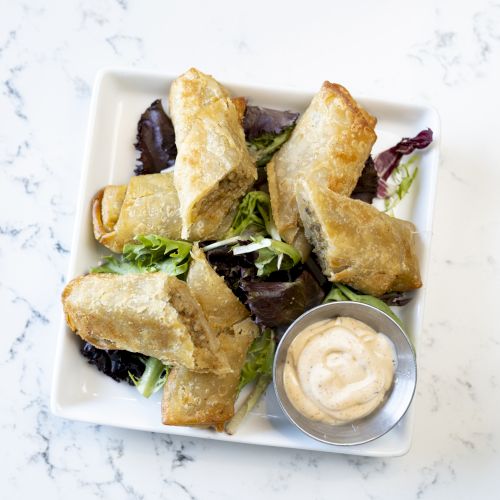 The image shows a plate of egg rolls on a bed of greens, accompanied by a dipping sauce in a small metal container.