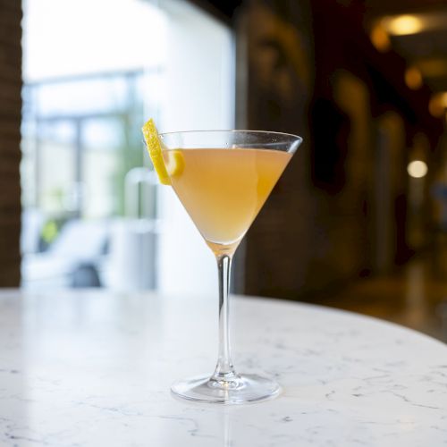 A cocktail in a martini glass garnished with a lemon twist sits on a marble table indoors, with a blurred background of a modern setting.