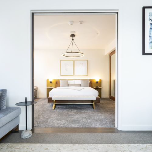 This image shows a modern bedroom with a white bed, side tables with lamps, a gray couch, a small table, and wall art.