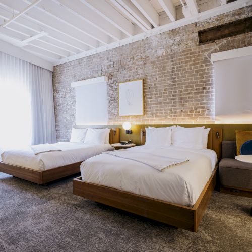 The image shows a modern hotel room with two beds, a couch, brick walls, white curtains, and stylish decor, creating a cozy atmosphere.