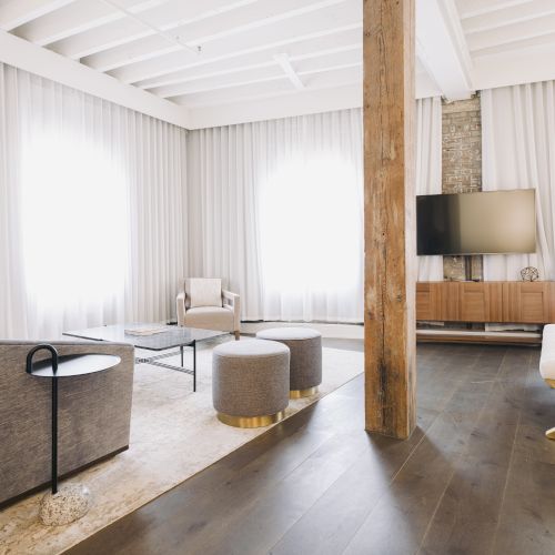 A modern living room with a sofa, armchair, stools, TV, and wooden accents on the floor and ceiling beams.