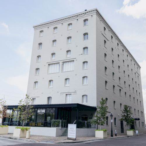 The image shows a tall, rectangular building with multiple windows and a sign out front. There are power lines and a clear blue sky in the background.