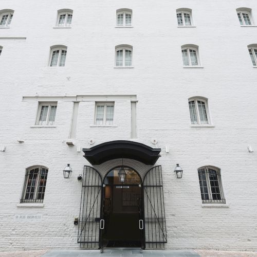 A white brick building with multiple windows and a black-arched entrance gate. The door is centered with lamps on either side of the entrance.