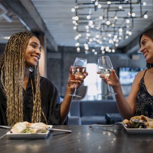 Two people are sitting at a table in a restaurant, smiling and clinking wine glasses, with plates of food in front of them.