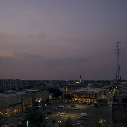 A cityscape at dusk with lit streets, buildings, few cars, and a distant church spire under a purple-gray sky, accompanied by a tall power line.