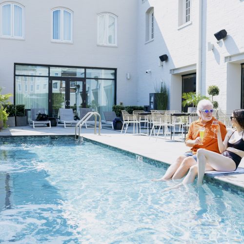 Two people are sitting by a pool, one wearing sunglasses and an orange outfit, and the other in a black swimsuit, enjoying a conversation.