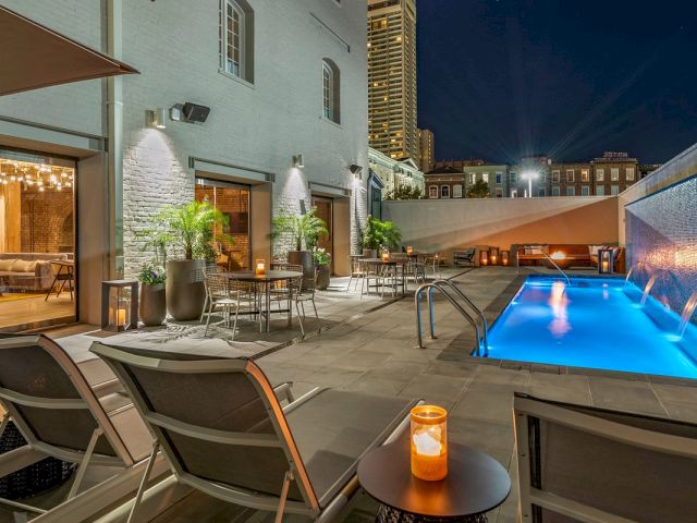 A nighttime view of a rooftop pool area with a lit pool, lounge chairs, tables, and plants.
