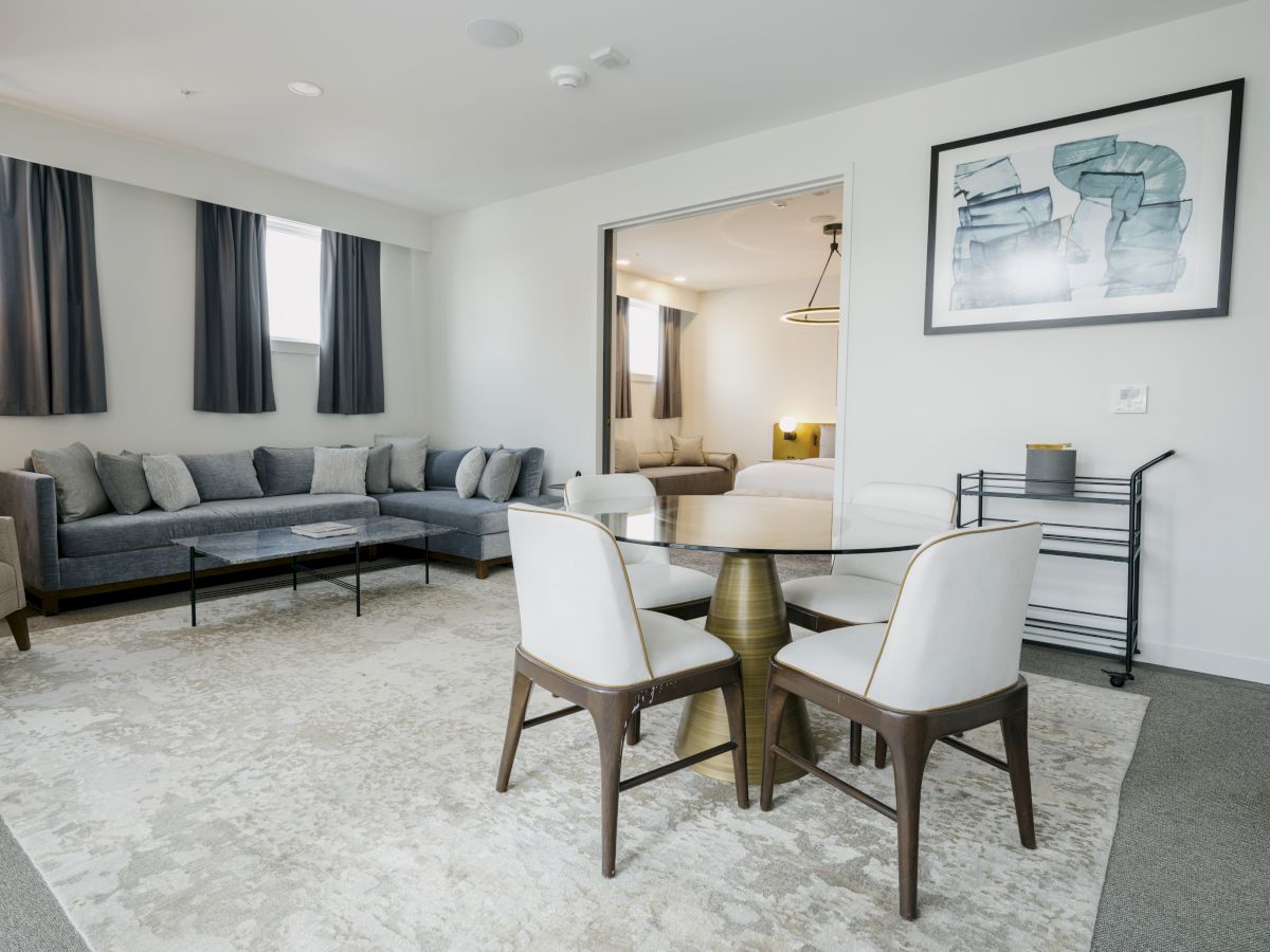 A modern living room features a gray sectional sofa, a round dining table with chairs, and a rug; a bedroom is visible through an open doorway.