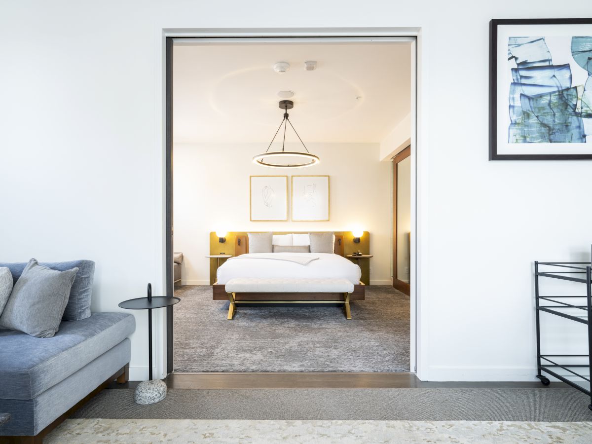 The image shows a modern bedroom with a neatly made bed, bedside tables, and a couch in the adjoining room, featuring minimalist decor.