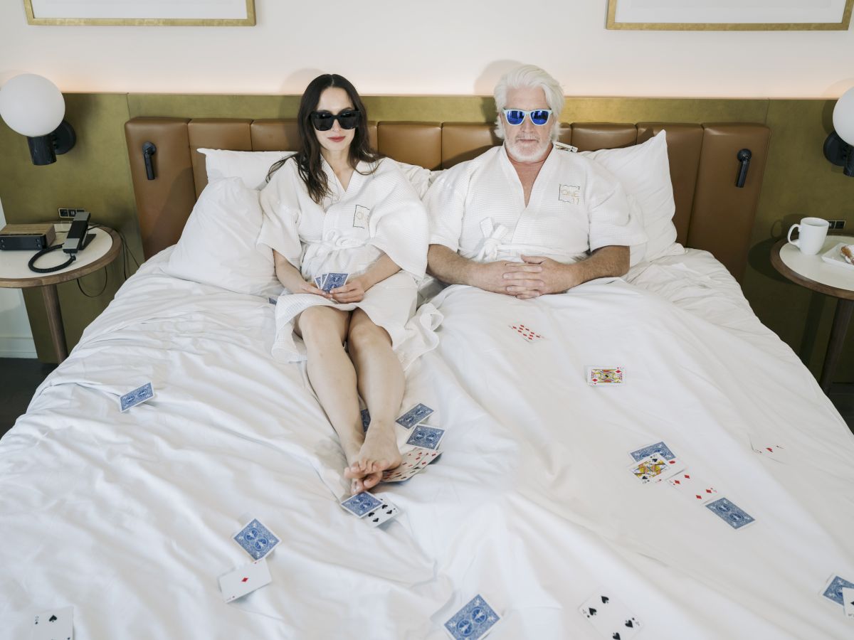 Two people wearing sunglasses and bathrobes are lying on a bed strewn with playing cards, in what appears to be a hotel room or bedroom.