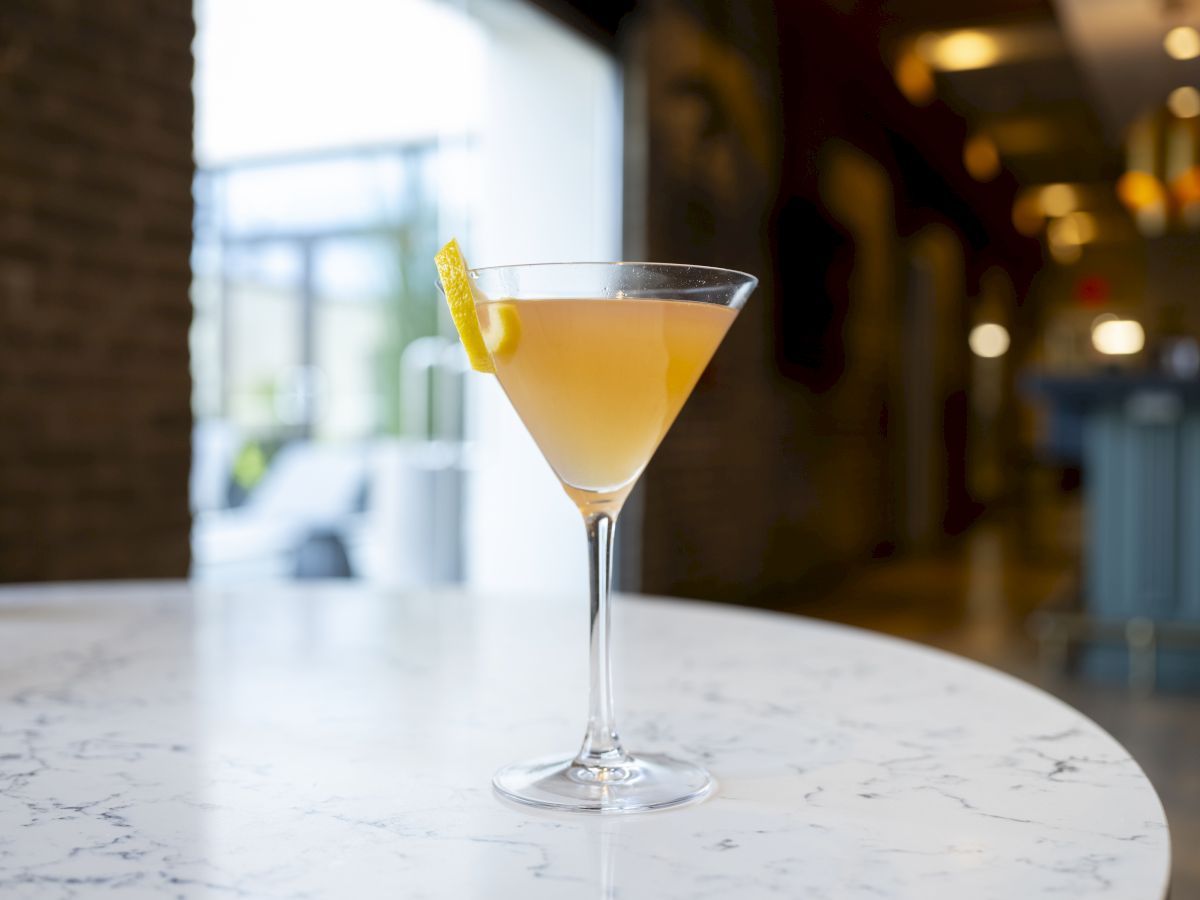 A cocktail in a stemmed glass with a lemon twist garnish placed on a marble table in a well-lit indoor setting.